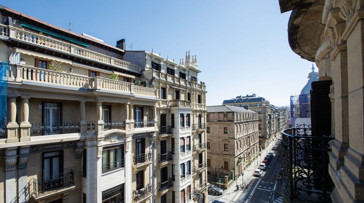 Vistas de la calle Prim desde un balcón en el Ensanche de San Sebastián (Donostia), mostrando fachadas históricas de piedra y balcones de hierro forjado bajo un cielo despejado.