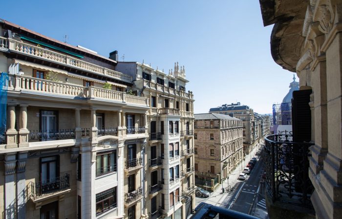 Vistas de la calle Prim desde un balcón en el Ensanche de San Sebastián (Donostia), mostrando fachadas históricas de piedra y balcones de hierro forjado bajo un cielo despejado.