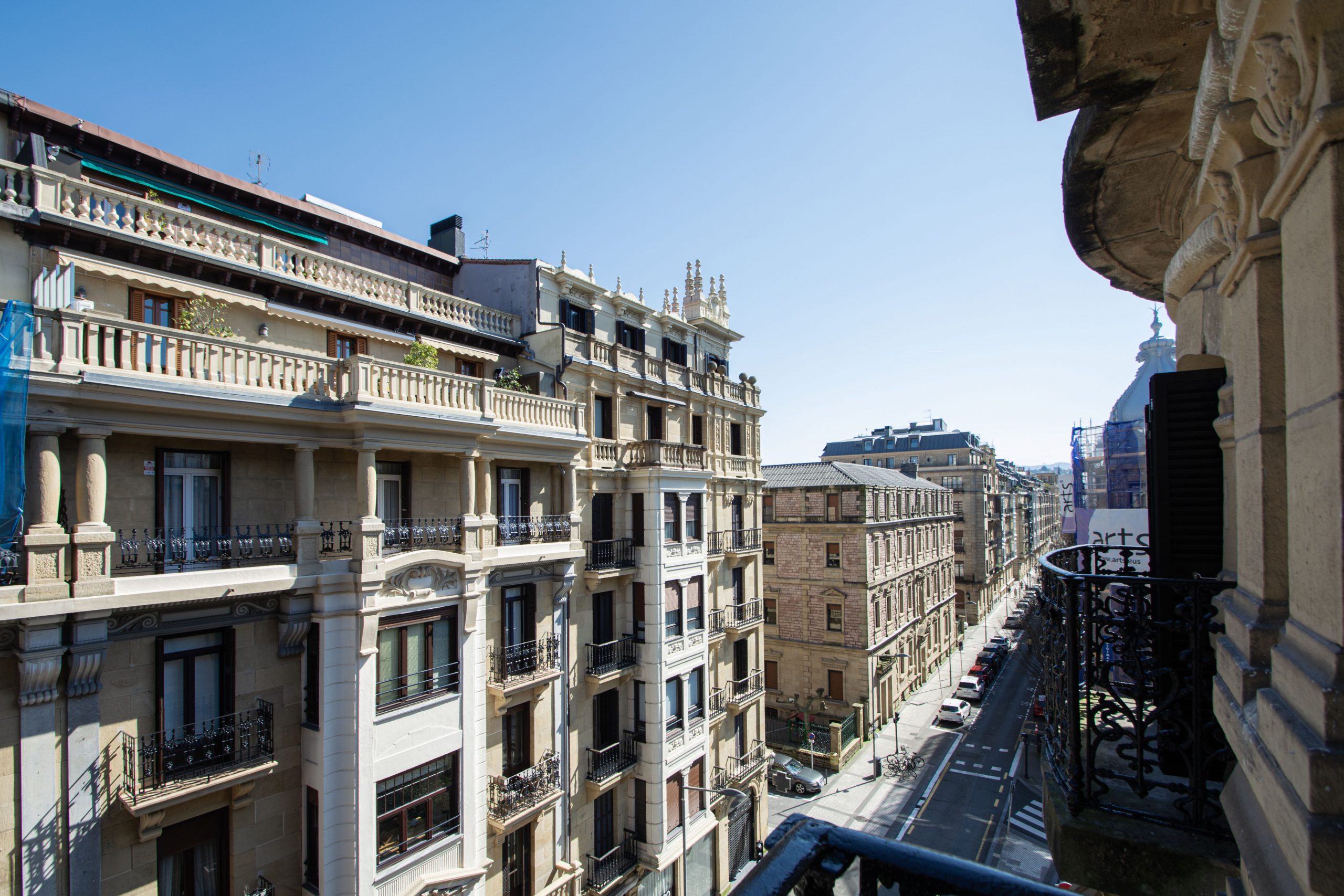 Vistas de la calle Prim desde un balcón en el Ensanche de San Sebastián (Donostia), mostrando fachadas históricas de piedra y balcones de hierro forjado bajo un cielo despejado.